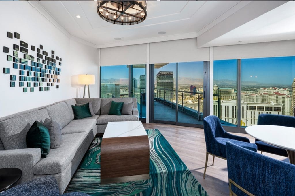 Modern living room with gray sectional sofa, teal accents, abstract wall art, large windows, city skyline view, and a round dining table with blue chairs.