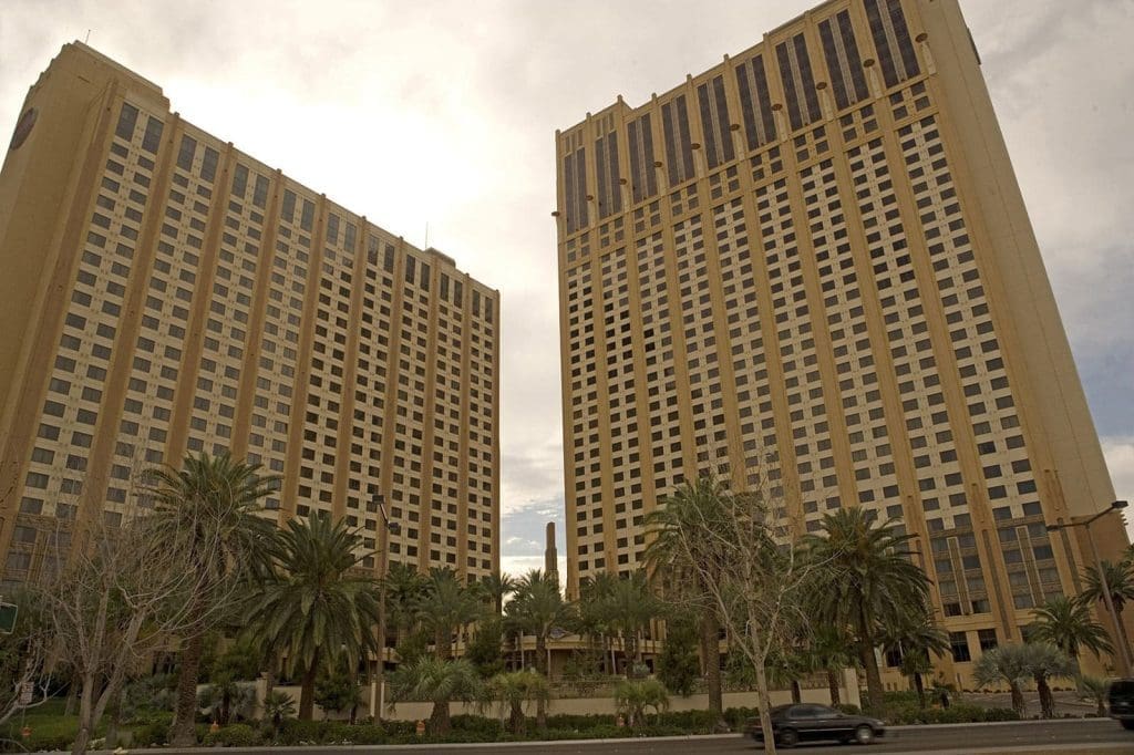 Two tall, beige high-rise buildings with vertical dark window columns stand side by side, surrounded by palm trees and a street with a passing car in front.