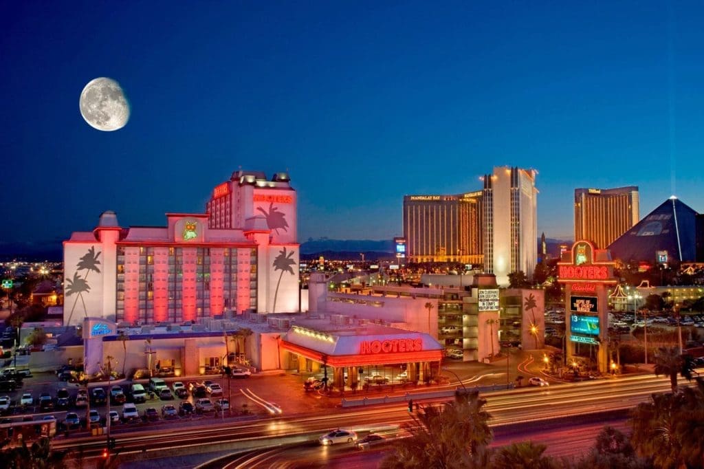 Night view of Las Vegas Strip with illuminated buildings, including a Hooters hotel and casino, traffic light trails, and a large moon visible in the sky.