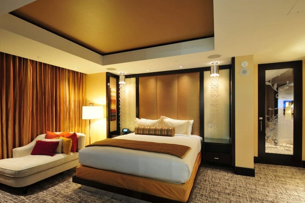 Modern hotel room with a king bed, padded headboard, chaise lounge, warm lighting, brown and gold accents, and floor-to-ceiling curtains.
