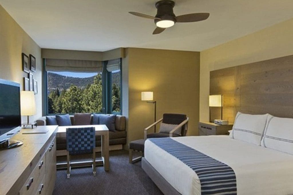 Modern hotel room with a large bed, desk, TV, armchair, sofa, and window overlooking trees and mountains. Neutral decor with blue accents.