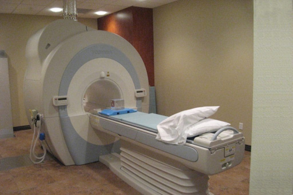 A medical MRI scanner with a patient table and pillow in an exam room with beige walls and a tiled floor.