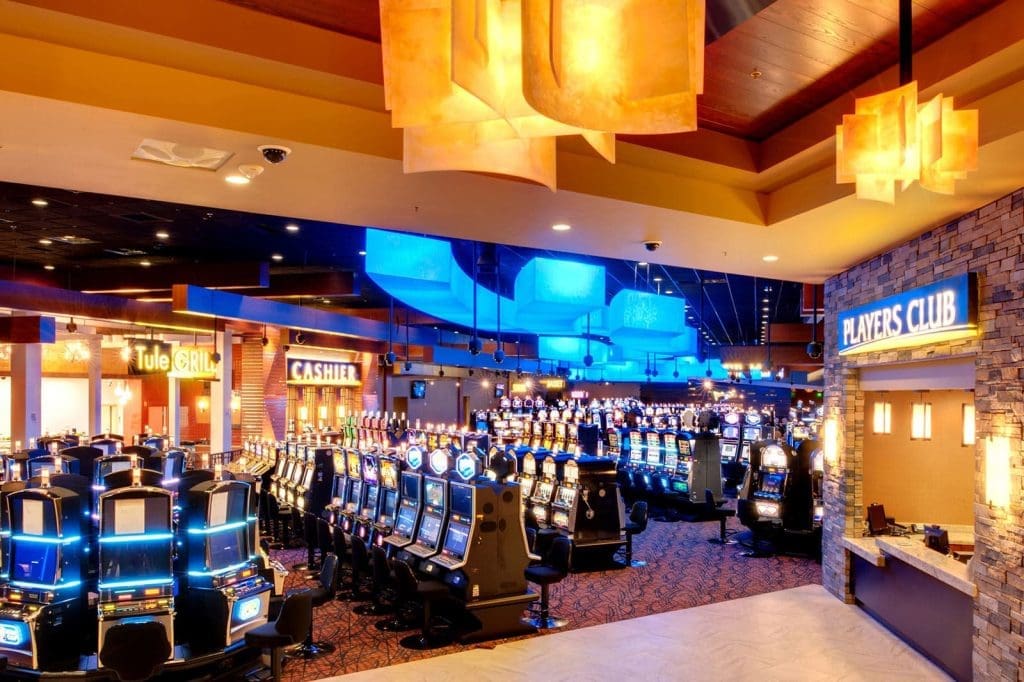 Casino interior with rows of illuminated slot machines, vibrant lighting, and signs for the Tule Grill, Cashier, and Players Club visible.