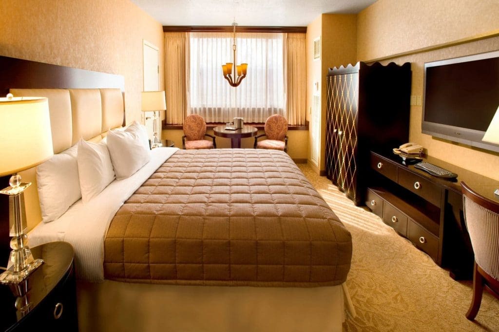 Hotel room with a large bed, bedside tables with lamps, a flat-screen TV on a dresser, a small table with two chairs, and a window with curtains.