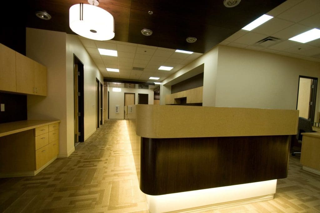 Reception desk in a modern office or medical building with wood and beige finishes, illuminated ceiling lights, and empty hallways.