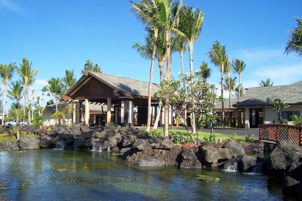 A tropical resort building with palm trees, a man-made pond, rocks, and small waterfalls under a clear blue sky.