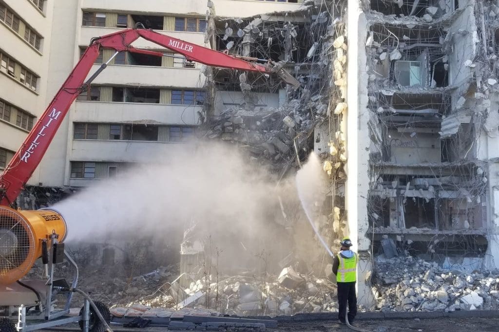 A worker sprays water to control dust while a building is being demolished with heavy machinery. Rubble and debris are visible on the ground.