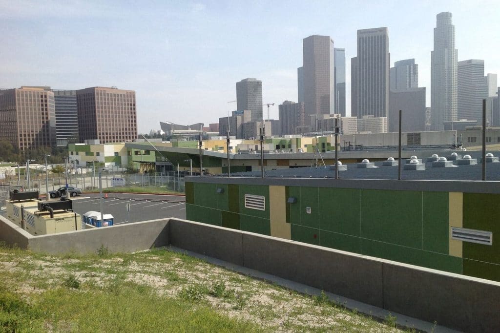 Buildings with green accents and an empty parking lot in the foreground, with the downtown Los Angeles skyline visible in the background on a clear day.