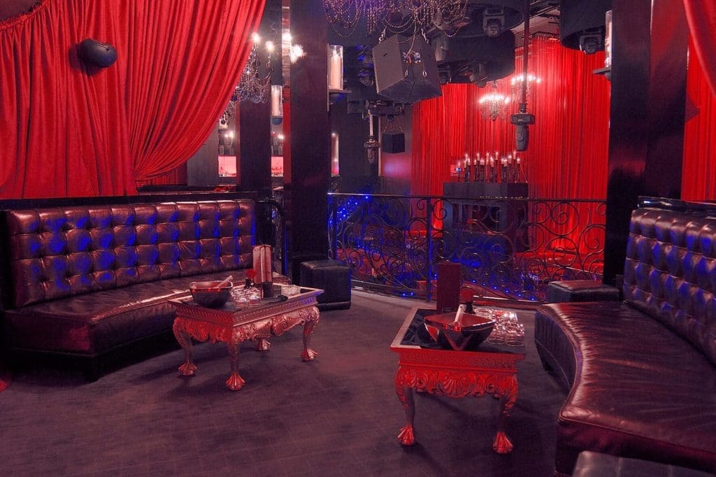 Luxurious nightclub interior with dark leather seating, ornate red tables, red curtains, chandeliers, and decorative railing.