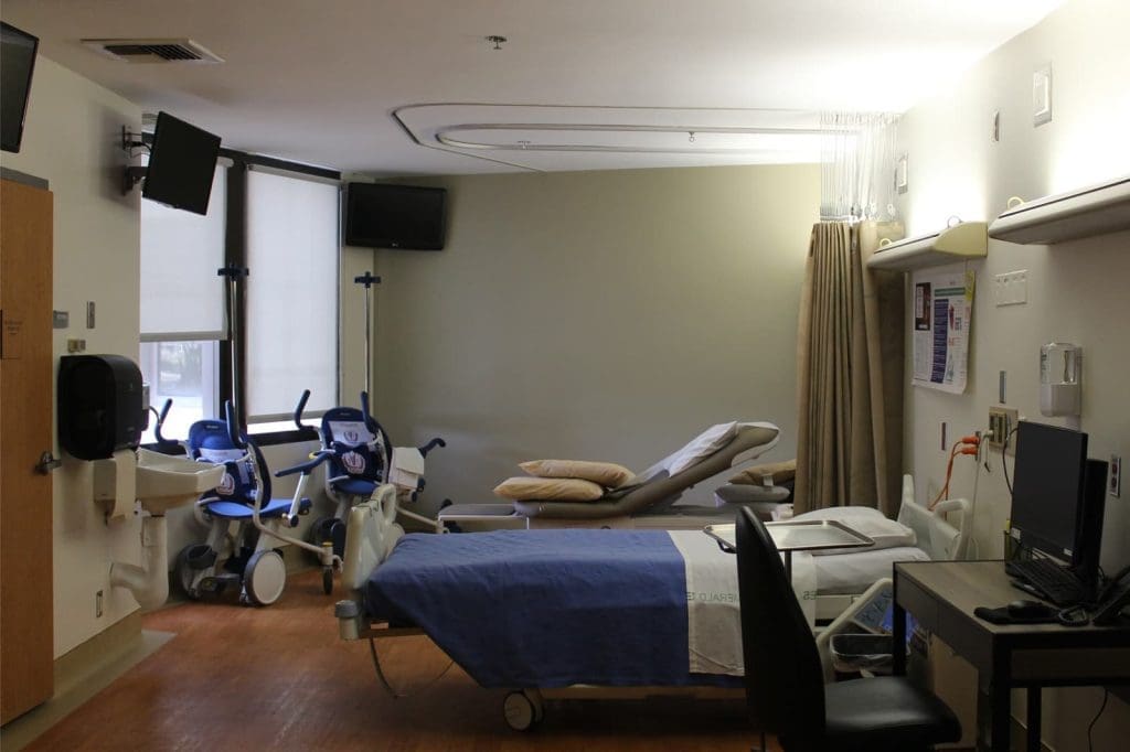 A hospital room with a bed, two chairs, medical equipment, a sink, a computer workstation, and wall-mounted monitors. The room is well-lit with natural and artificial light.