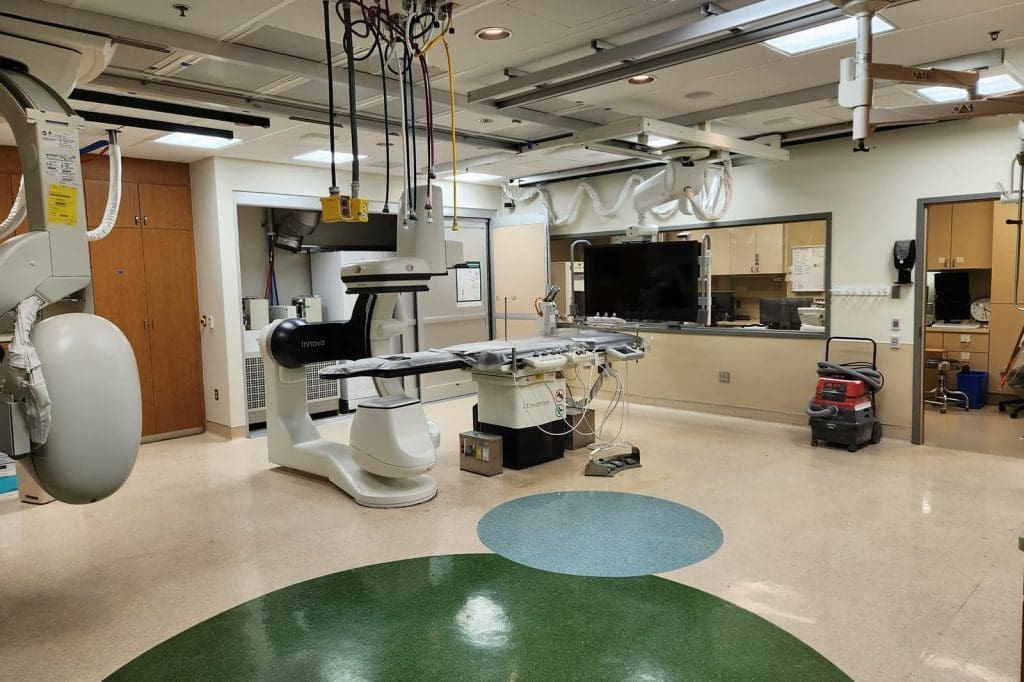A hospital procedure room with medical imaging equipment, a patient table, monitors, cabinets, and a floor cleaning machine in the corner.