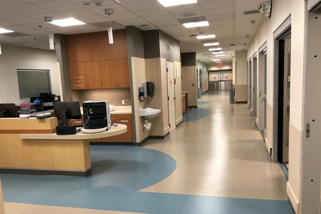 A clean, modern hospital hallway with patient rooms on the right, a nurses’ station on the left, and overhead fluorescent lighting.