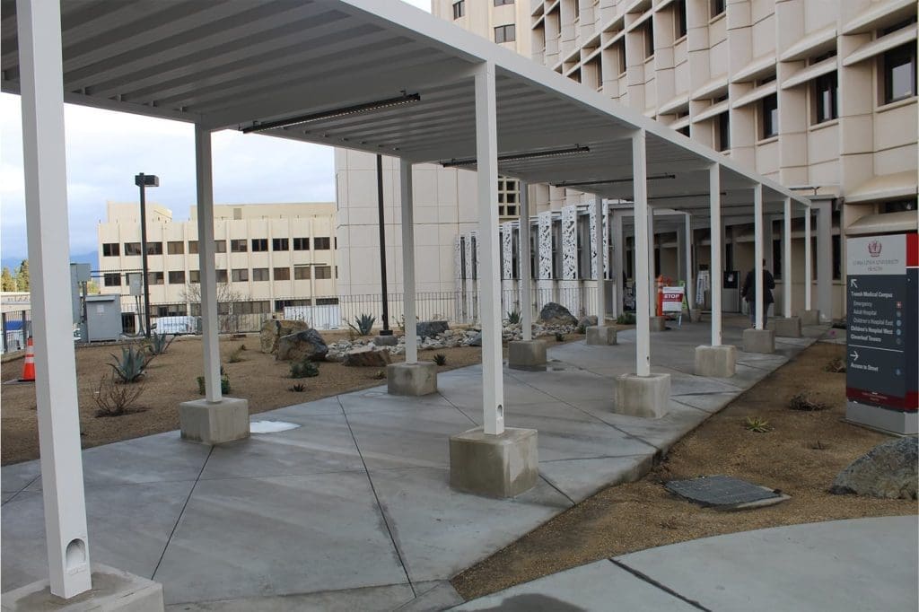 Covered walkway with white metal supports outside a multi-story building, surrounded by dry landscaping and signage.