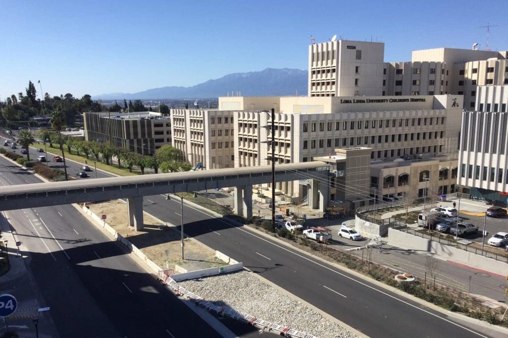 A hospital complex with multiple beige buildings and a skybridge over a wide road, with mountains visible in the background under a clear blue sky.