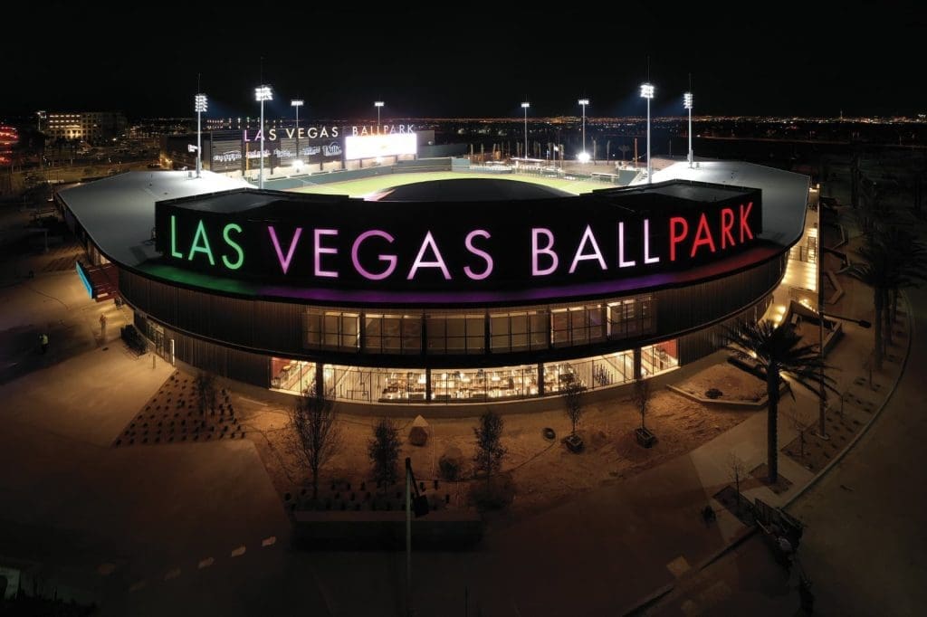 Aerial night view of Las Vegas Ballpark with illuminated signage and stadium lights, surrounded by an empty parking area.