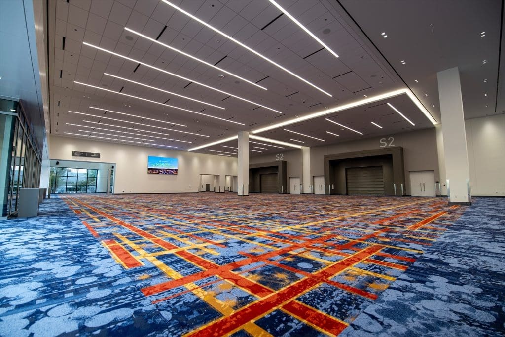 Spacious convention center hall with patterned carpet, high ceilings, bright lighting, and large doors labeled S2. A digital screen displays an image near glass doors.