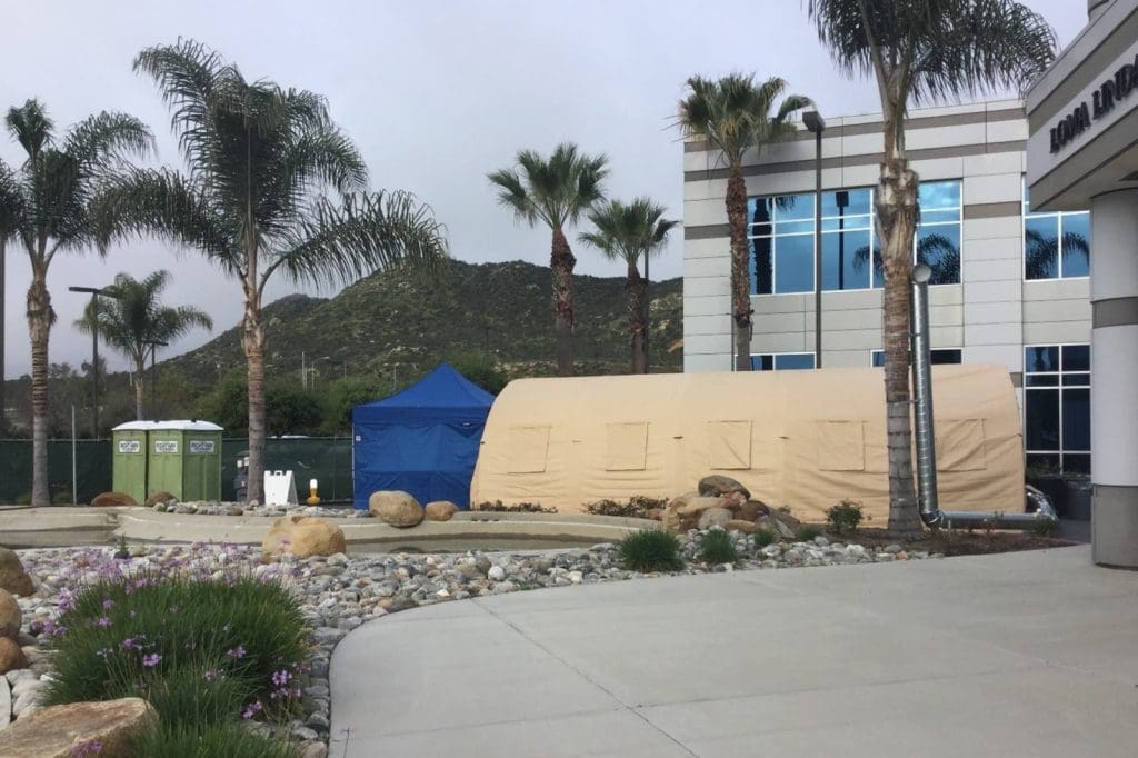 Large beige tent and blue canopy set up beside a modern building with palm trees, mountains in the background, and portable toilets nearby.