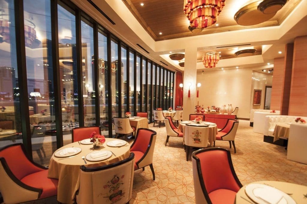 Elegant restaurant interior with round tables, red cushioned chairs, large floor-to-ceiling windows, decorative lighting, and a view of city buildings outside.