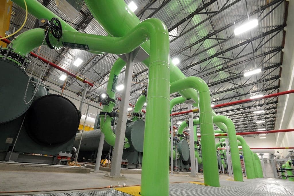 Large green industrial pipes run through a facility with metal grating floors, overhead lighting, and machinery in the background.