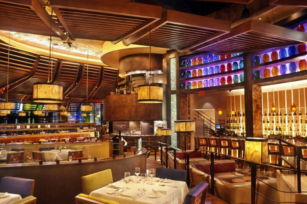 Modern restaurant interior featuring wooden accents, pendant lighting, colorful glass decorations on shelves, set tables, and upholstered seating areas.