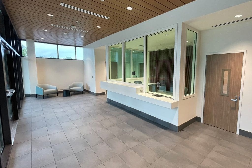 Modern waiting area with two chairs, a small table, tiled floor, wood-paneled ceiling, large windows, and a counter with glass partition next to a closed wooden door.