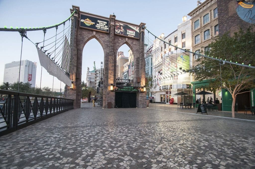 A stone arch bridge with overhead signs stands at the entrance to a pedestrian plaza lined with shops and buildings, under a clear sky.