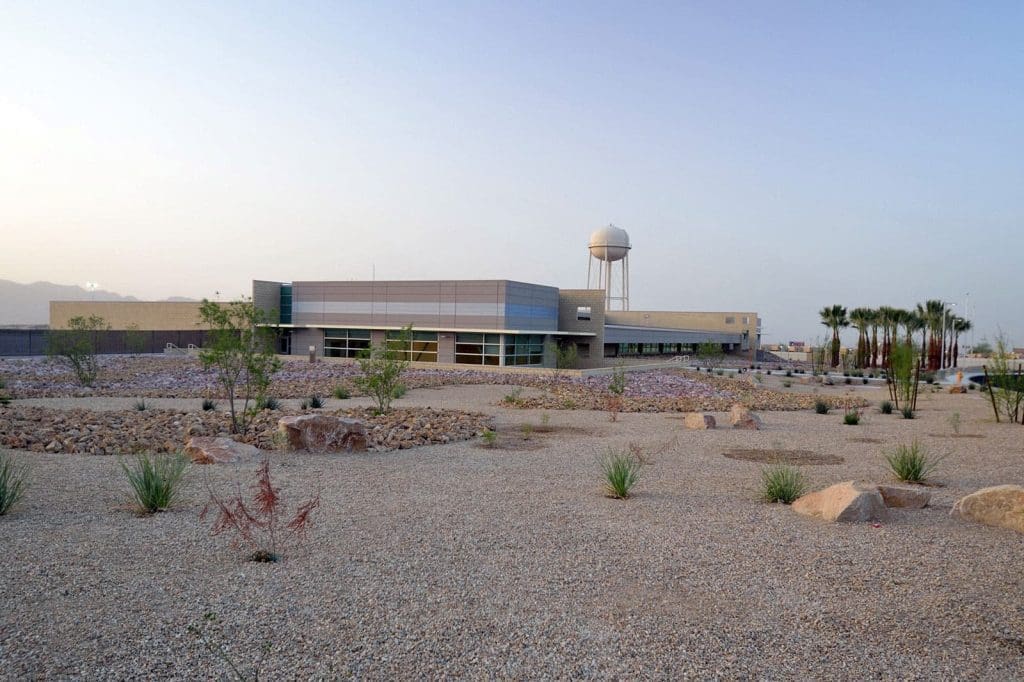 A modern low-rise building with large windows stands in a desert landscape with gravel, rocks, sparse plants, and a water tower in the background.