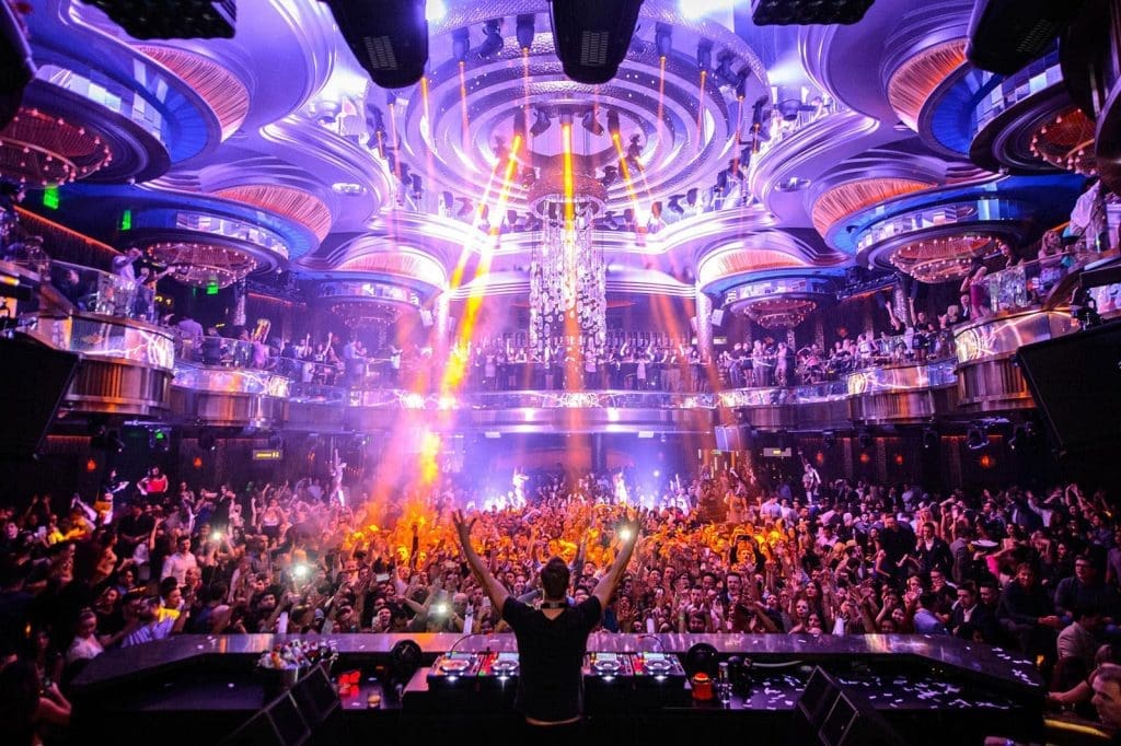 A DJ performs on stage in a crowded nightclub with colorful lights, lasers, and a large cheering audience.