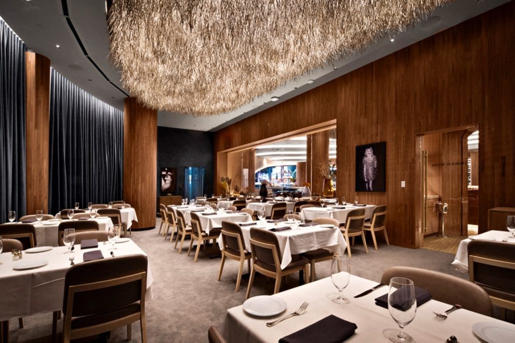 Modern restaurant dining room with wooden walls, white tablecloths, neatly set tables, upholstered chairs, and a large textured light fixture hanging from the ceiling.