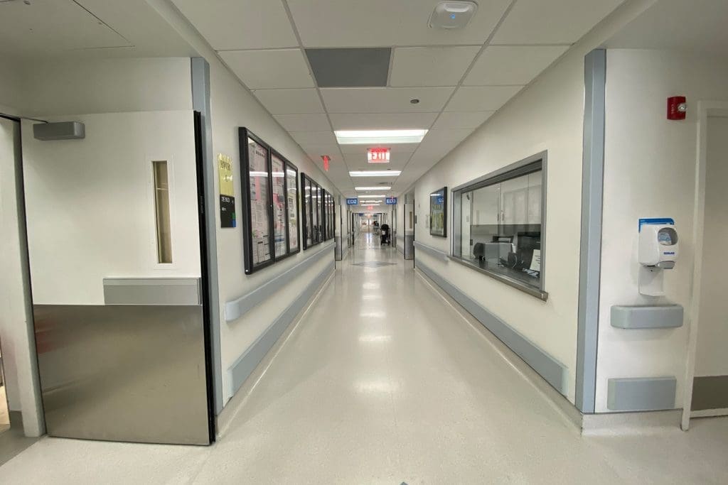 A brightly lit hospital hallway with white walls, bulletin boards on the left, a window on the right, and an open door at the far end.