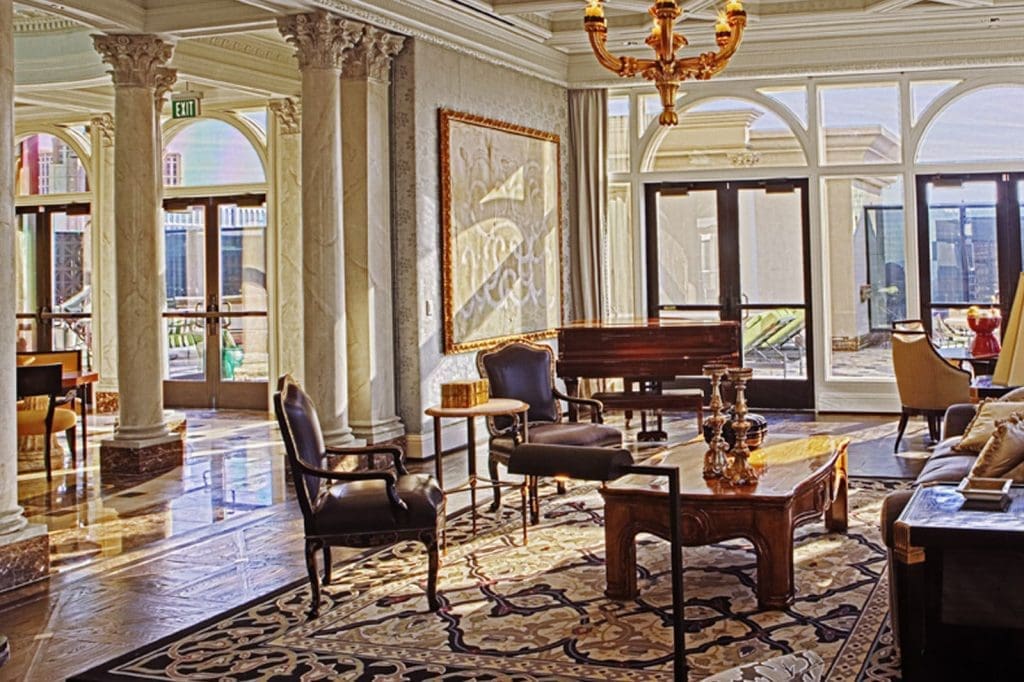 Elegant hotel lobby with marble columns, large windows, classic furniture, ornate rug, and a chandelier, lit by natural sunlight.
