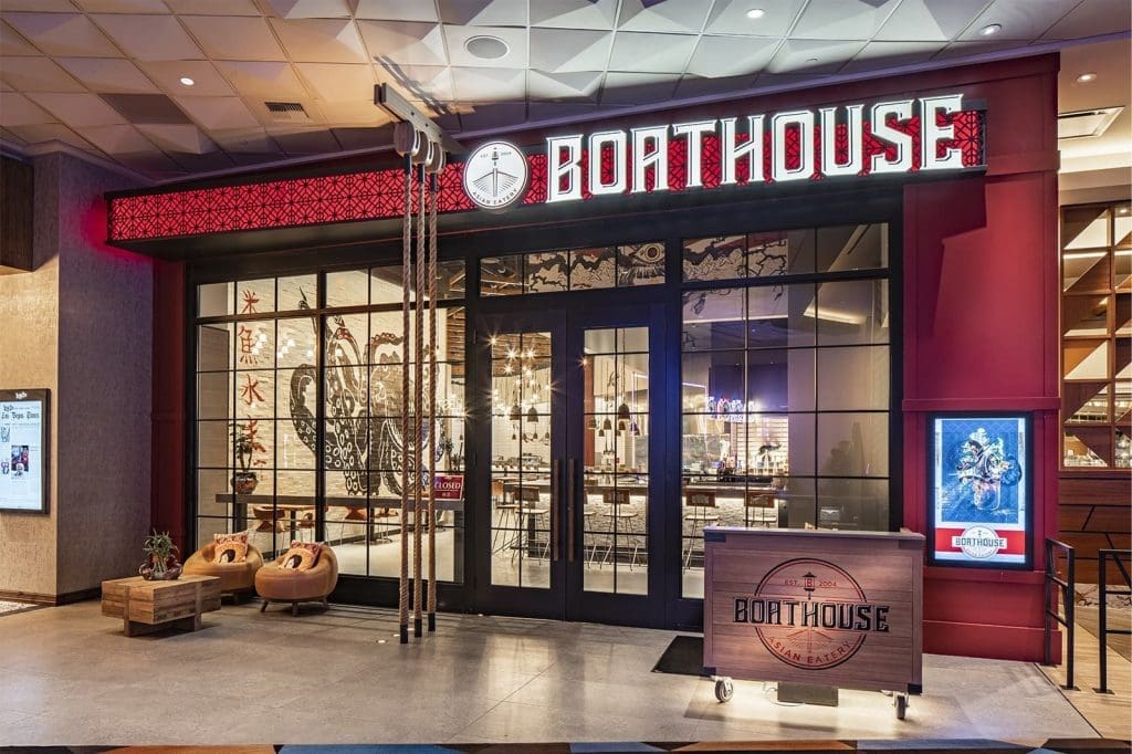 Exterior view of Boathouse restaurant with large illuminated sign, decorative nautical elements, glass doors, and indoor seating visible through the windows.