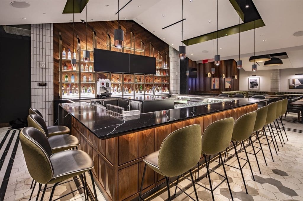 Modern bar with a black marble countertop, green upholstered barstools, and shelves of liquor bottles behind the counter, under pendant lights in a stylish, well-lit room.