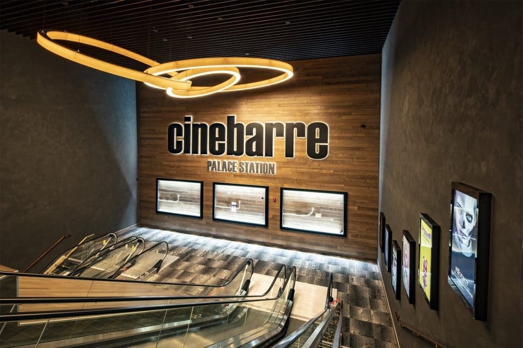 A modern escalator leads to the entrance of cinebarre Palace Station, with movie posters displayed on wood-paneled and gray walls.