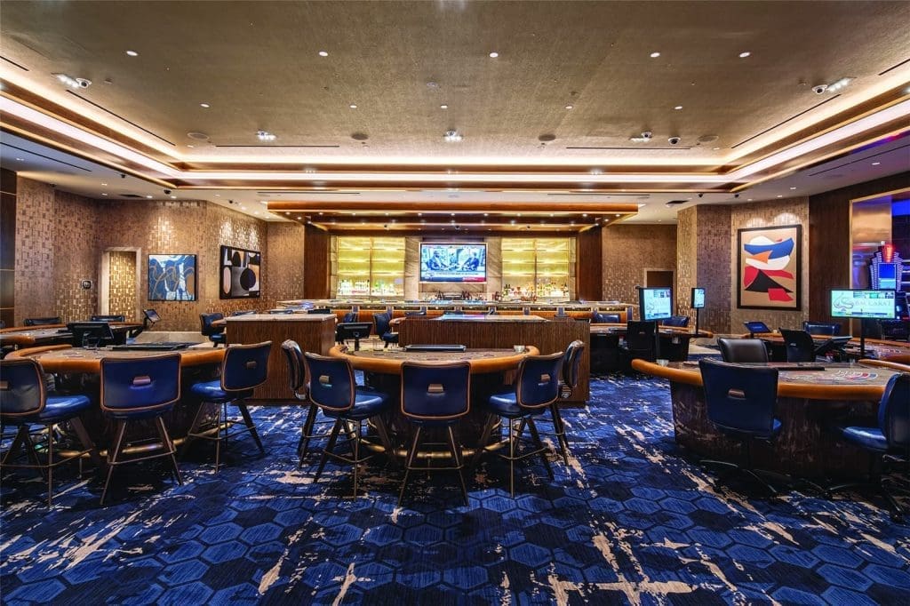 Modern casino interior with empty gaming tables, blue patterned carpet, decorative lighting, and artwork on the walls. A bar and television are visible at the back of the room.