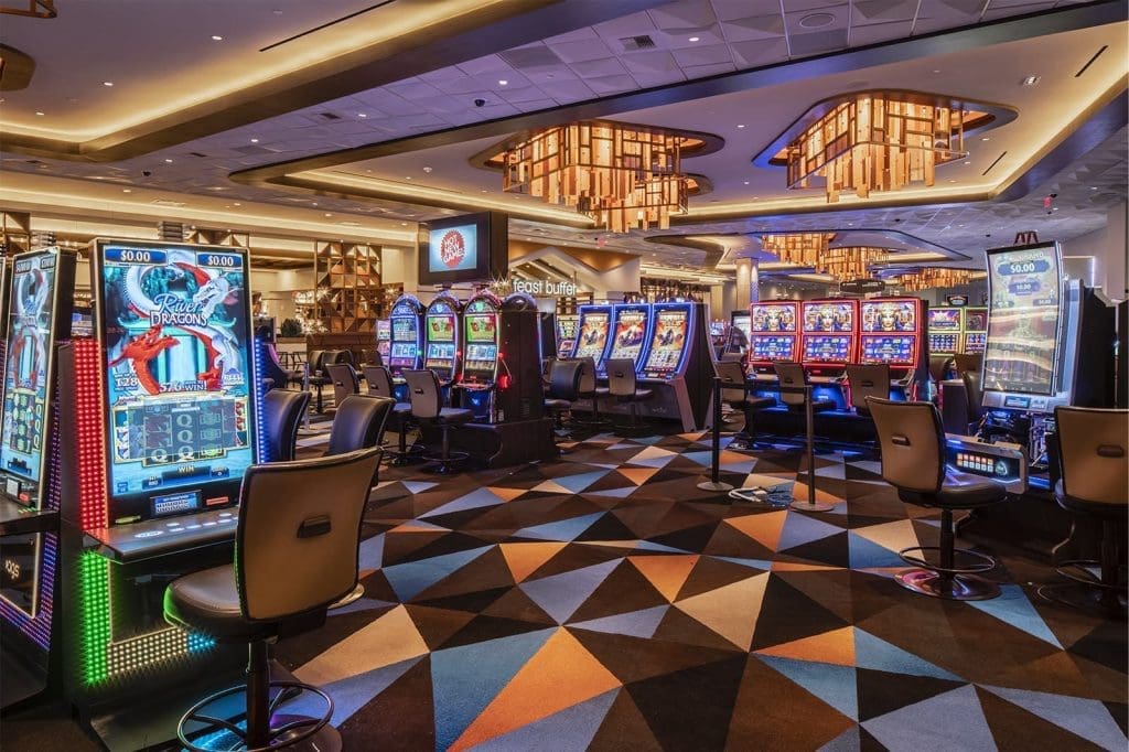 Casino interior with rows of colorful slot machines, patterned carpet, modern lighting fixtures, and empty chairs, creating a lively gaming atmosphere.