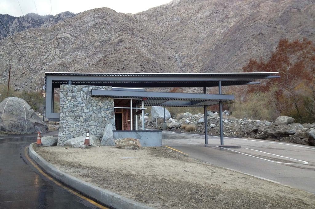 A modern stone and metal toll booth stands beside a curved road, with mountains and rocky terrain in the background.