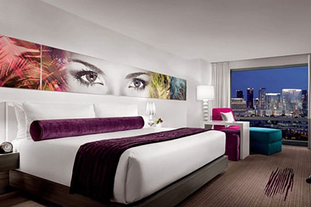 Modern hotel room with a large bed, purple accents, wall art of eyes above the headboard, seating area, and a window view of a city skyline at night.