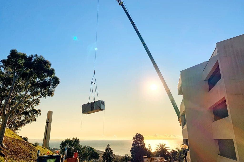 A crane lifts a large metal object near a building at sunset, with trees and the ocean visible in the background.