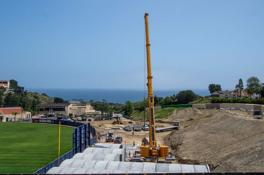 A large yellow crane operates at a construction site near a grassy field, with the ocean and several buildings visible in the background under a clear sky.