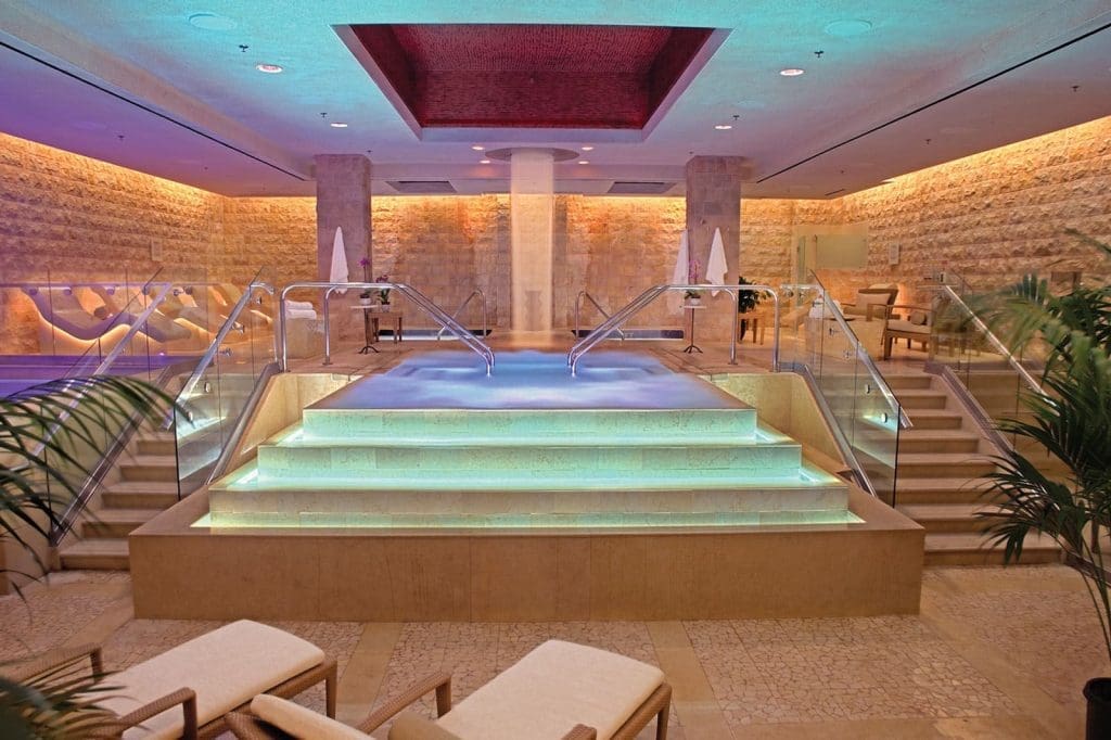 Indoor spa area with illuminated central pool, stairs with glass railings, stone walls, and ambient colored lighting; lounge chairs and plants in the foreground.