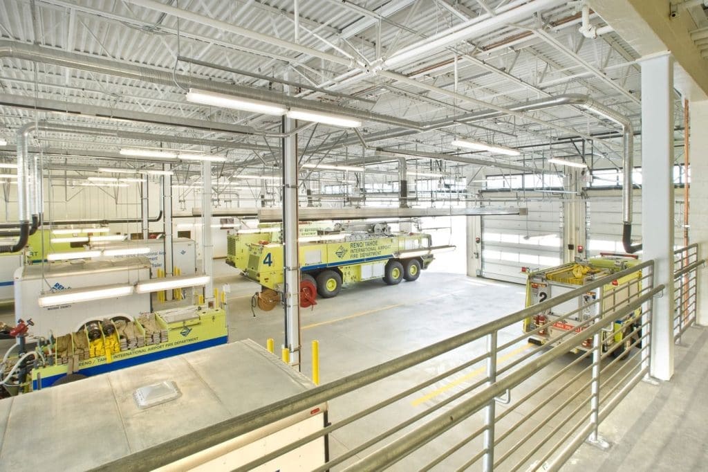 Interior view of a bright, spacious fire station bay with multiple large yellow fire trucks parked inside, seen from an upper walkway.
