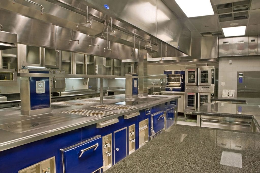 A modern commercial kitchen with stainless steel surfaces, blue cabinetry, multiple ovens, and industrial appliances under bright overhead lighting.