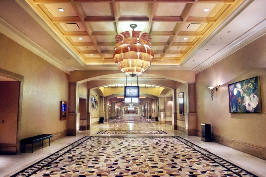 Wide hotel hallway with patterned floor, ornate ceiling light fixture, wall art, benches, and a digital sign suspended from the ceiling at the center.