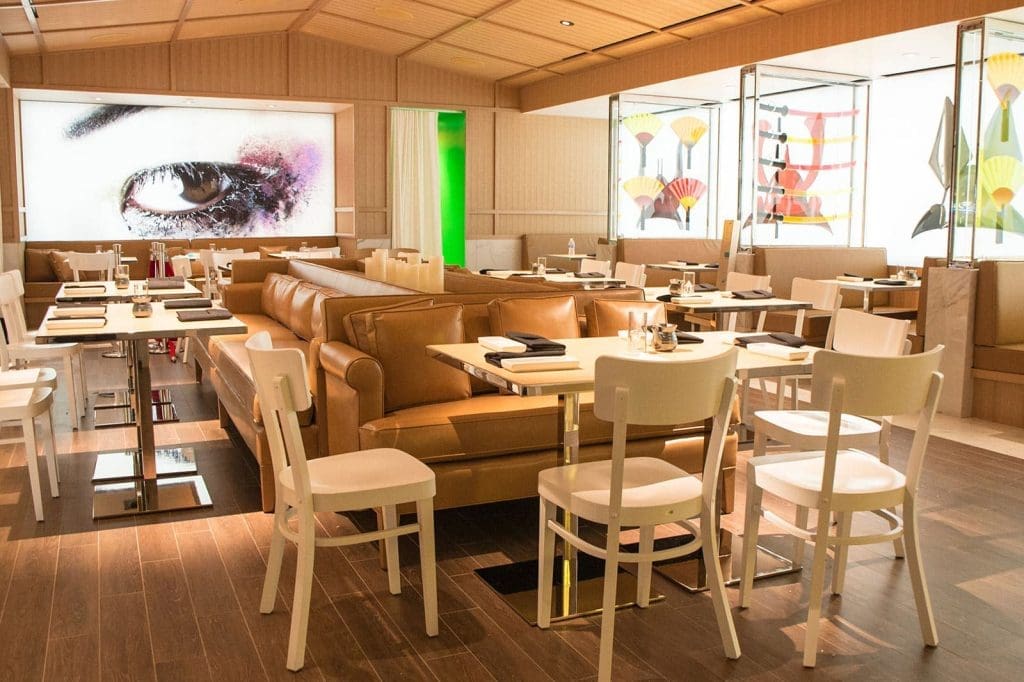 Modern restaurant interior with tan leather couches, white chairs, set tables, wood flooring, and large contemporary artwork on the walls.