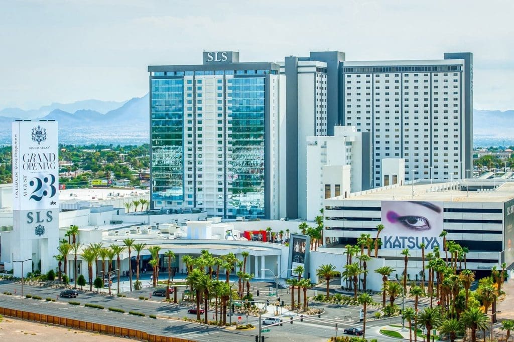 A view of the SLS hotel and casino complex in Las Vegas, featuring modern high-rise buildings, palm trees, and billboards, with mountains visible in the background.