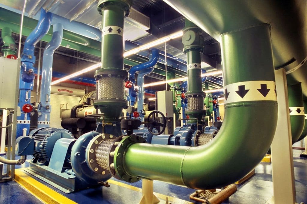 Large industrial green and blue pipes with directional arrow labels, mechanical equipment, and valves inside a brightly lit facility.