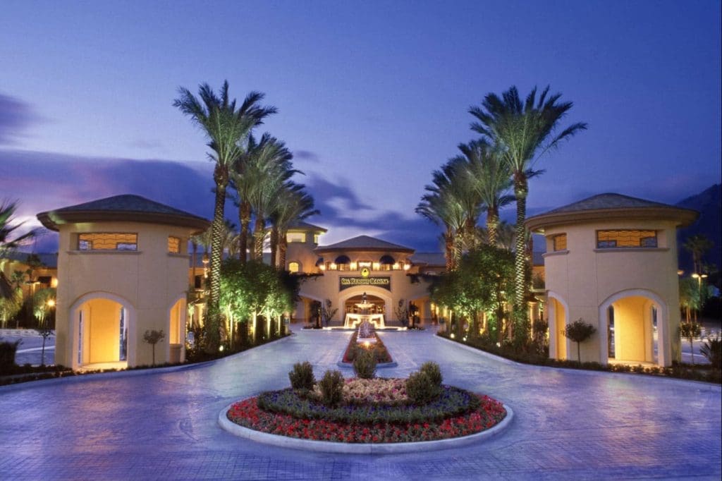 A luxury resort entrance with palm trees, landscaped gardens, round pavilions, and illuminated pathways at dusk.