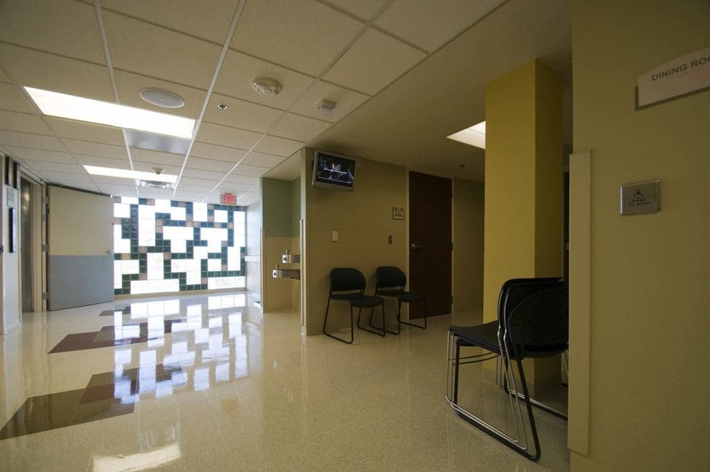 Empty hallway in a medical facility with chairs along the wall, a television mounted near the ceiling, and patterned glass windows at the end of the corridor.