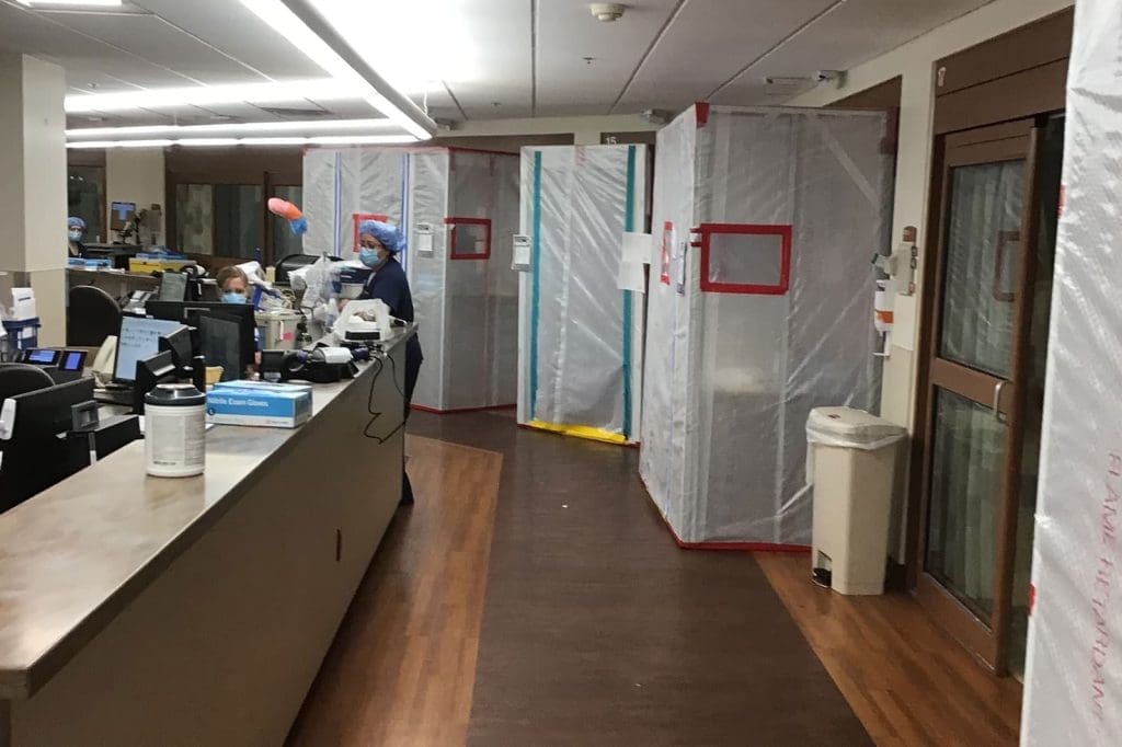 Hospital hallway with plastic containment barriers and healthcare workers at desks, likely indicating infection control or construction in progress.